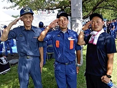 ポンプ操法大会で消防団の仲間と敬礼する私（左）。一番身近な共同体を平和にしようと「まぶる」精神で活動した。まぶるは地元の方言で、優しく見守るという意味だ