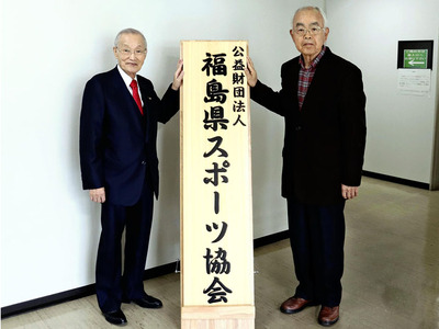 県体育協会から改称した「県スポーツ協会」の看板を手にする須佐喜夫会長と副会長の私（右）。一層の競技力向上や競技の普及に努めたい＝４月